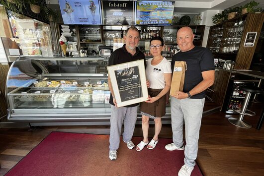 Markus Zlatnik, Irene Jandl und Bürgermeister Georg Rosner stehen nebeneinander im Lokal. Dahinter ist die Eisvitrine zu sehen.