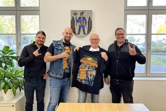 Vier Mitglieder des Rock Kollektiv Süd und der Bürgermeister stehen nebeneinander. Der Bürgermeister hält ein T-Shirt in der Hand und präsentiert es in die Kamera.